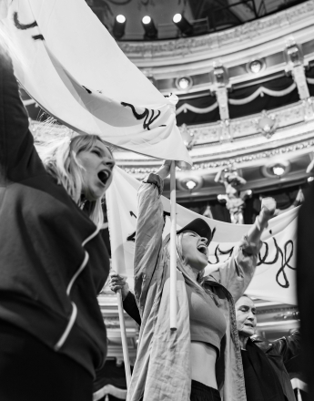 Czarno-białe zdjęcie. Na pierwszym planie Karolina Kamińska i Karolina Kazoń we wnętrzu teatru. Aktorki trzymają baner na długich drążkach, unoszą ręce i krzyczą, scena wygląda jak protest. W tle widoczne są balkony, kolumny i oświetlenie przestrzeni teatralnej.